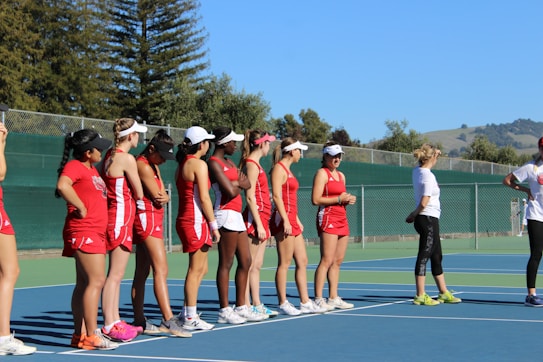 A group of women in red athletic uniforms and white visors stand in a line on a tennis court. They appear to be listening to a person dressed in casual athletic wear, who is standing to the right of the group. The background includes a green fence and trees, as well as distant hills under a clear blue sky.