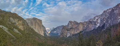 A panoramic view of a lush green valley surrounded by towering rocky cliffs and mountains, with a partly cloudy sky overhead. Some portions of the mountains are covered with patches of snow, indicating a cold climate.