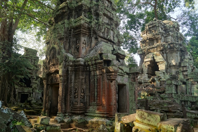 Ancient stone temple ruins surrounded by lush green foliage. The structures are intricately carved and appear weathered, with parts of the architecture in a state of disrepair. Large trees with dense canopies add to the scene, casting dappled shadows over the ruins.