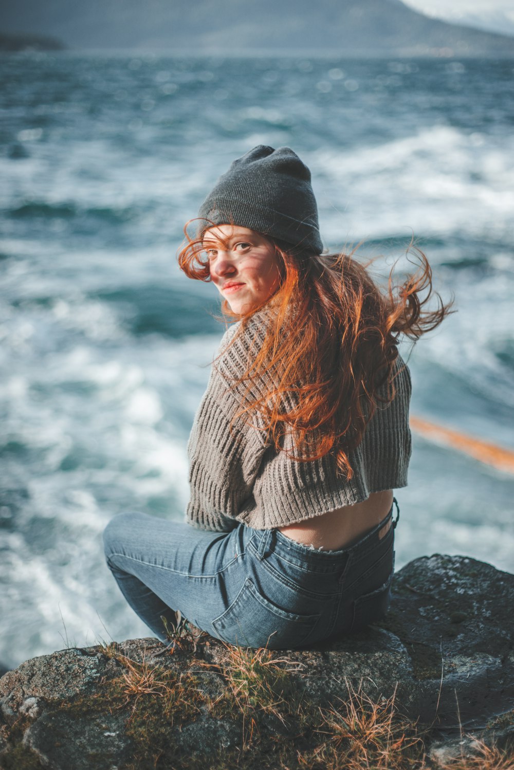 woman in brown knit sweater and blue denim jeans sitting on rock near sea during daytime