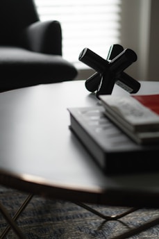 A modern, minimalist setting featuring a round table with a smooth surface. On the table, there are a few neatly stacked books and a black decorative object resembling a jack. In the background, a part of a dark, upholstered chair and a softly lit window with blinds can be seen.