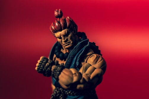 A muscular action figure with red hair tied in a top knot and wearing a blue gi poses against a vibrant red background. The figure's expression is intense, with focused eyes and clenched fists wrapped in rope.