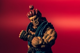A muscular action figure with red hair tied in a top knot and wearing a blue gi poses against a vibrant red background. The figure's expression is intense, with focused eyes and clenched fists wrapped in rope.