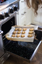 A warm kitchen scene showing a smiling teenage baker holding a tray of freshly baked chocolate chip cookies.