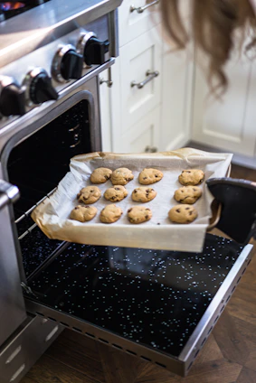 A warm kitchen scene showing a smiling teenage baker holding a tray of freshly baked chocolate chip cookies.