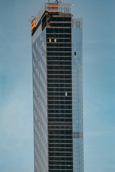 A tall, modern skyscraper with a sleek and minimalist design, featuring a combination of reflective glass panels and dark, vertical stripes. The upper section is under construction, visible with scaffolding and an orange safety netting. The building is set against a clear, pale blue sky.