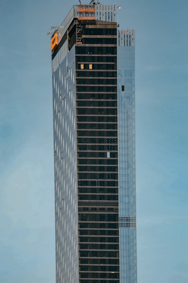 A tall, modern skyscraper with a sleek and minimalist design, featuring a combination of reflective glass panels and dark, vertical stripes. The upper section is under construction, visible with scaffolding and an orange safety netting. The building is set against a clear, pale blue sky.
