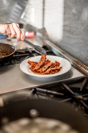 A hand is using black tongs to place strips of crispy cooked bacon onto a white plate. The plate is situated on a metal stove, and in the background, a blurred kitchen environment is visible.