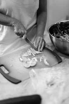 A person is slicing vegetables on a cutting board with a knife, wearing a light-colored shirt. Nearby, there is a metal bowl containing chopped greens, likely on a kitchen countertop.