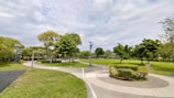 A well-maintained park with a curving pathway surrounded by lush green grass and trees. The sky is partly cloudy, and there are several blue solar-powered streetlights lining the path. Neatly trimmed bushes and small garden areas enhance the landscape.