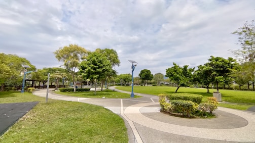 A well-maintained park with a curving pathway surrounded by lush green grass and trees. The sky is partly cloudy, and there are several blue solar-powered streetlights lining the path. Neatly trimmed bushes and small garden areas enhance the landscape.