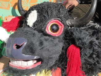 A detailed and colorful bull mask with exaggerated features, including large eyes, prominent teeth, and bright decorative elements such as horns and fabric accents. The mask is placed on a vibrant patterned cloth.