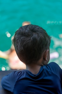 A child is seen from behind, leaning forward on a railing. The child is wearing a dark blue shirt and appears to be looking at someone swimming in a body of water on a bright, sunny day. The water is a bright turquoise color, with visible white reflections from the sunlight.