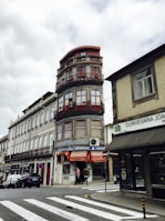 A tall, narrow building with rounded corners and red window frames stands at a street intersection. The ground floor features orange awnings and signs for Casa Lourenço, a shop selling cheeses and meats. There are people walking by on the cobblestone pavement, and a second building is adjacent, showcasing tiled patterns and an awning over stores. Cars are parked along the street, accompanied by traffic signs.