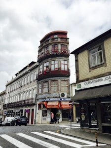 A tall, narrow building with rounded corners and red window frames stands at a street intersection. The ground floor features orange awnings and signs for Casa Lourenço, a shop selling cheeses and meats. There are people walking by on the cobblestone pavement, and a second building is adjacent, showcasing tiled patterns and an awning over stores. Cars are parked along the street, accompanied by traffic signs.