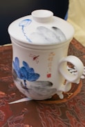 A ceramic mug with a lid, featuring intricate blue floral designs and black accents. There is calligraphy and possibly a red stamp on the side. The mug is placed on a richly patterned surface.