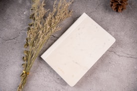 An overhead view of a white marble notebook lying on a textured gray surface. A bundle of dried grasses and a pine cone are placed beside it, creating a natural, minimalist composition.