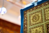 A close-up view of a patterned carpet hanging with a clip, featuring intricate designs in shades of yellow, blue, and burgundy. The carpet has a floral motif enclosed in square sections, and its texture appears plush. The background is softly blurred, with hints of colorful lights.