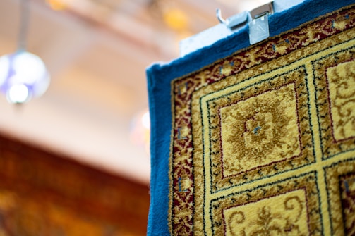 A close-up view of a patterned carpet hanging with a clip, featuring intricate designs in shades of yellow, blue, and burgundy. The carpet has a floral motif enclosed in square sections, and its texture appears plush. The background is softly blurred, with hints of colorful lights.