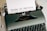 A vintage typewriter with a sheet of paper inserted, displaying the text 'Terms of Service'. The typewriter has a dark green body and black keys, with a brand name on the top right side.