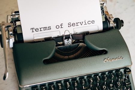 A vintage typewriter with a sheet of paper inserted, displaying the text 'Terms of Service'. The typewriter has a dark green body and black keys, with a brand name on the top right side.