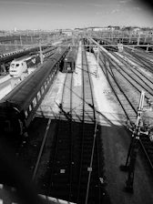 A black and white photograph of a train yard with multiple train tracks and several stationary trains. The tracks extend into the distance, converging towards the horizon. Overhead wires and poles run parallel to the tracks, contributing to the complex, industrial atmosphere. Buildings and infrastructure can be seen in the background under a clear sky.