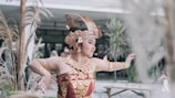 A dancer wearing traditional ornate attire and a headpiece performs a graceful pose in an outdoor setting with plants in the background.