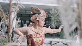A dancer wearing traditional ornate attire and a headpiece performs a graceful pose in an outdoor setting with plants in the background.