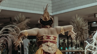 A person wearing an elaborate traditional costume with ornate patterns and headgear is captured from behind, performing a graceful dance. The setting seems to be outdoors with decorative plants and a modern structure in the background.