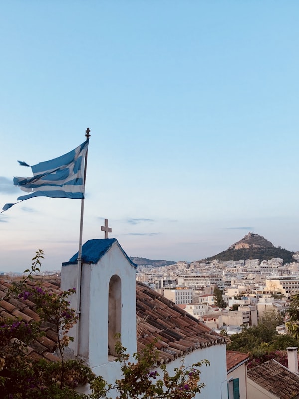 Greek flag flying over a whitewashed chapel with Mount Lycabettus behind
