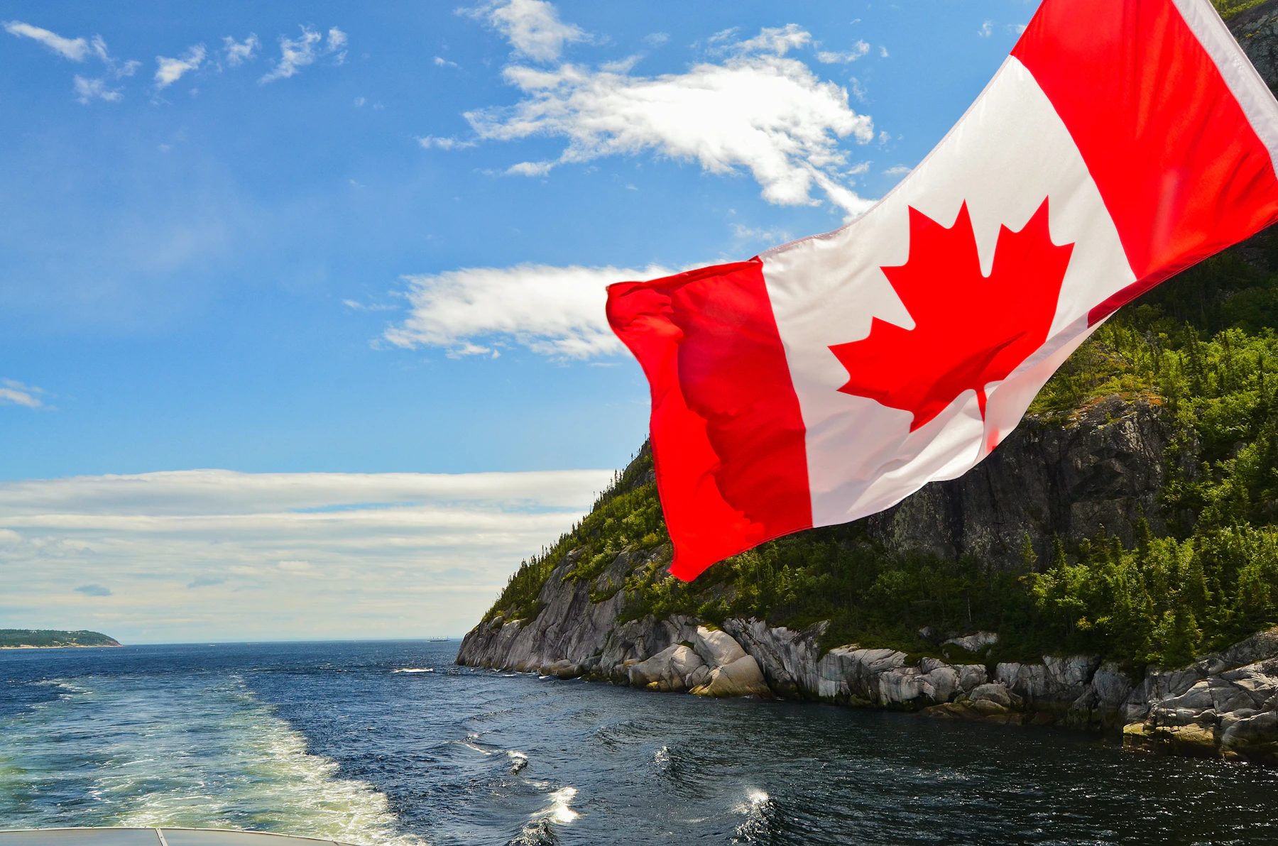 A Canadian flag stands beside rock formations above water.