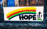 A colorful banner displayed on a metal fence in front of a brick building. The banner features a rainbow with horizontal stripes of yellow, red, green, and blue. Below the rainbow, the word 'HOPE' is written in bold black letters. To the right of the word, there are three black crosses on green bases illustrated.