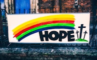 A colorful banner displayed on a metal fence in front of a brick building. The banner features a rainbow with horizontal stripes of yellow, red, green, and blue. Below the rainbow, the word 'HOPE' is written in bold black letters. To the right of the word, there are three black crosses on green bases illustrated.
