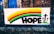A colorful banner displayed on a metal fence in front of a brick building. The banner features a rainbow with horizontal stripes of yellow, red, green, and blue. Below the rainbow, the word 'HOPE' is written in bold black letters. To the right of the word, there are three black crosses on green bases illustrated.
