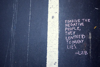 Text written in pink chalk on a dark asphalt surface conveys a message about forgiving negative people. The text reads: 'FORGIVE THE NEGATIVE PEOPLE, THEY LEARNED TO MANY LIES. -LiB'. The message is placed adjacent to a white line that runs vertically on the ground, which is cracked and weathered.