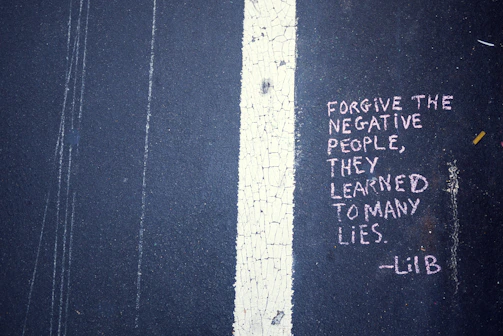 Text written in pink chalk on a dark asphalt surface conveys a message about forgiving negative people. The text reads: 'FORGIVE THE NEGATIVE PEOPLE, THEY LEARNED TO MANY LIES. -LiB'. The message is placed adjacent to a white line that runs vertically on the ground, which is cracked and weathered.