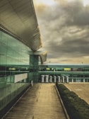 A modern architectural building featuring a large, sleek roof with metal and glass elements. The structure extends into the distance, showcasing a series of metal chimneys or vents. The facade is made up of reflective glass panels, and the ground is a wooden deck or walkway. The sky is filled with dramatic, overcast clouds, suggesting an impending storm.