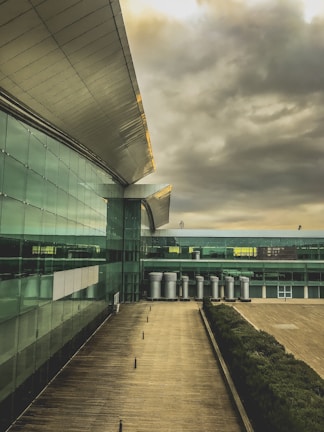 A modern architectural building featuring a large, sleek roof with metal and glass elements. The structure extends into the distance, showcasing a series of metal chimneys or vents. The facade is made up of reflective glass panels, and the ground is a wooden deck or walkway. The sky is filled with dramatic, overcast clouds, suggesting an impending storm.