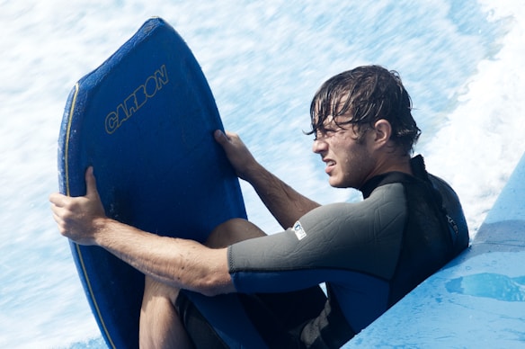 A person gripping a blue bodyboard with intense focus and determination. The individual is wearing a wetsuit, with water splashing around, suggesting an aquatic or surfing activity.