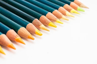 A row of colored pencils is arranged diagonally, featuring sharp points and displaying a gradient of colors from orange to green. The pencils have dark green barrels and are set against a plain white background.