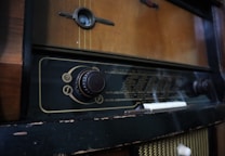 A vintage radio with a wooden exterior and a large tuning dial on the front. The radio display is marked with various frequency numbers and has an old-fashioned design. The exterior shows signs of wear, with some chipping and fading.
