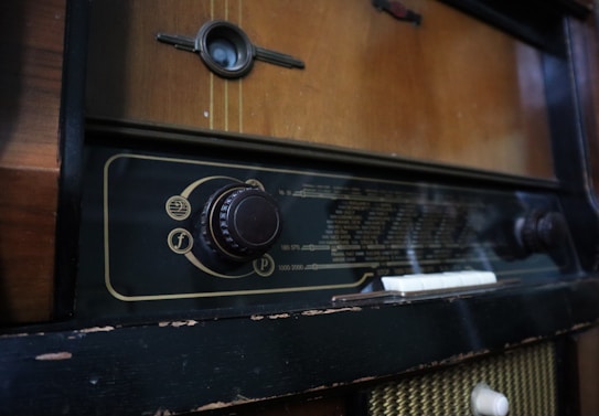 A vintage radio with a wooden exterior and a large tuning dial on the front. The radio display is marked with various frequency numbers and has an old-fashioned design. The exterior shows signs of wear, with some chipping and fading.