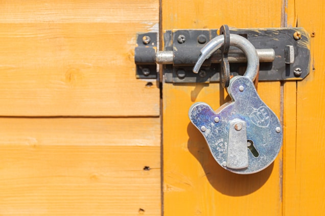 A weathered metallic padlock hangs on a closed latch fixed to a wooden surface. The wood is painted a bright yellow-orange, highlighting the lock's silvery, aged texture.