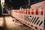 A nighttime construction site features bright red and white barriers lined along a dirt path. There are various construction materials and signs visible, with buildings illuminated by artificial lights in the background.