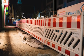 A nighttime construction site features bright red and white barriers lined along a dirt path. There are various construction materials and signs visible, with buildings illuminated by artificial lights in the background.