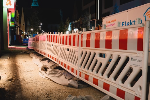 A nighttime construction site features bright red and white barriers lined along a dirt path. There are various construction materials and signs visible, with buildings illuminated by artificial lights in the background.