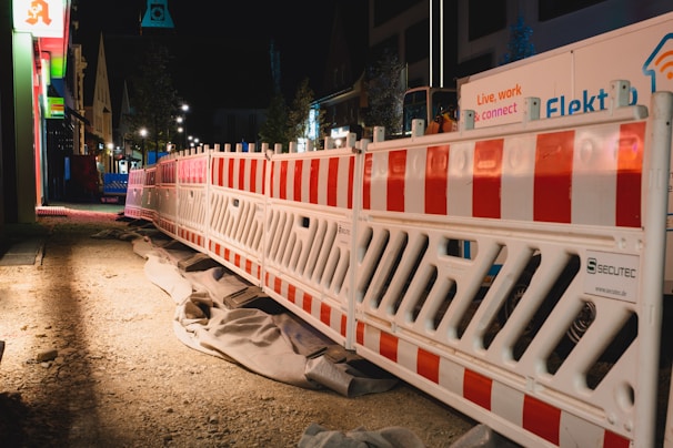 A nighttime construction site features bright red and white barriers lined along a dirt path. There are various construction materials and signs visible, with buildings illuminated by artificial lights in the background.