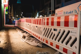 A nighttime construction site features bright red and white barriers lined along a dirt path. There are various construction materials and signs visible, with buildings illuminated by artificial lights in the background.