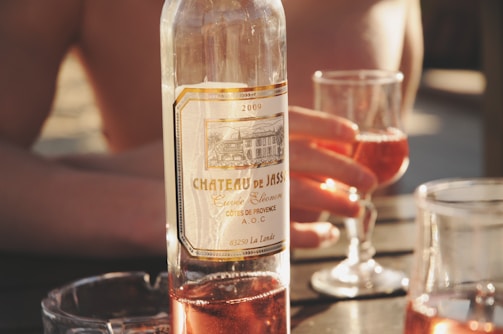 A wine bottle labeled 'Chateau de Jass' is placed on a table. Two glasses filled with rosé wine are visible, one being held by a hand. The setting appears relaxed and sunlit, suggesting a casual outdoor gathering.