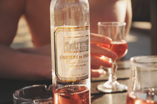 A wine bottle labeled 'Chateau de Jass' is placed on a table. Two glasses filled with ros&eacute; wine are visible, one being held by a hand. The setting appears relaxed and sunlit, suggesting a casual outdoor gathering.